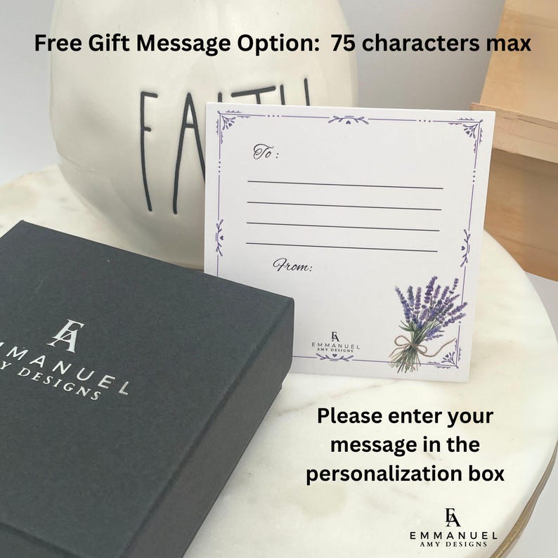 May include: A white rectangular gift card with a purple floral border, a lavender bouquet illustration, and the text To: and From: with lines for writing. A black gift box with the white logo EMMANUEL AMY DESIGNS is positioned next to the card.