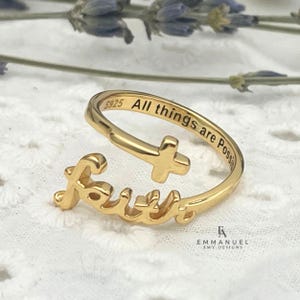 May include: A gold-colored ring with the word "Faith" in cursive script. The ring features a cross design and the inscription "All things are Possible." The ring is on a white lace surface.
