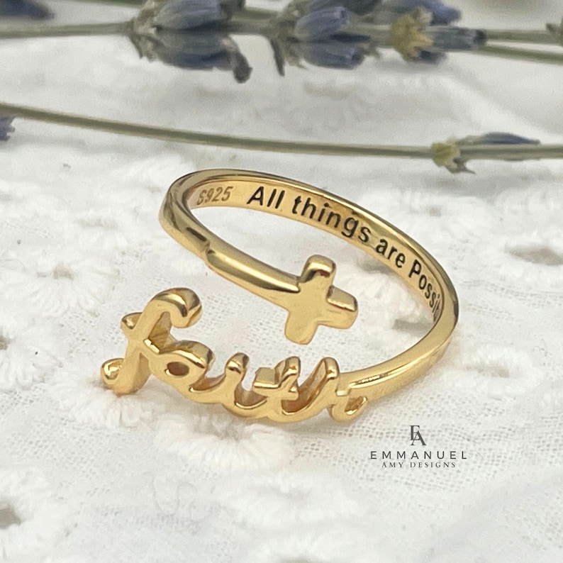 A gold-colored ring with the word "Faith" in cursive script. The ring features a cross design and the inscription "All things are Possible." The ring is on a white lace surface.