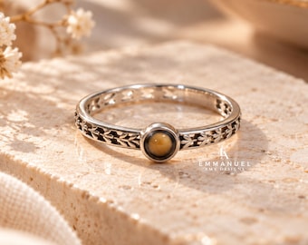 May include: A silver-toned ring featuring a central round tiger's eye gemstone and a band decorated with a repeating leaf pattern. The ring is engraved with the text EMMANUEL AMY DESIGNS near the setting. The metal has a polished finish.