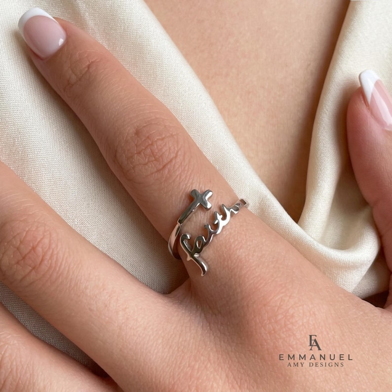 Peut inclure: Une bague ouverte de couleur argent&eacute;e pr&eacute;sentant le mot faith &eacute;crit en lettres cursives, avec un petit symbole de croix int&eacute;gr&eacute; au design. La bague poss&egrave;de une finition m&eacute;tallique polie et porte l'inscription EMMANUEL AMY DESIGNS visible.