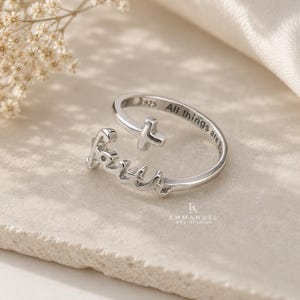 Peut inclure: Une bague ouverte de couleur argent&eacute;e pr&eacute;sentant le mot Faith en &eacute;criture cursive et un petit symbole de croix. La bande int&eacute;rieure est grav&eacute;e avec le texte S925 et All things are possible. La bague a une finition m&eacute;tallique polie et r&eacute;glable.