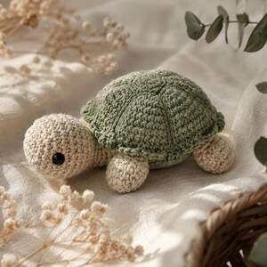Peut inclure: Une tortue en crochet avec une carapace texturée vert sauge et une tête, un cou et des membres de couleur crème. La tortue possède un petit œil noir rond et est réalisée avec des points de laine visibles, formant un motif de carapace arrondi.