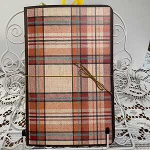 May include: A brown notebook with a plaid cover in shades of brown, orange, and blue. The notebook is closed and has a gold ribbon tied around it.