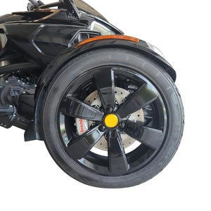 May include: Close-up of a black three-wheeled motorcycle. The front wheel has a black rim with a yellow center cap. The tire is black with the brand name "KENDA" visible. The brake caliper is red with the "brembo" logo.