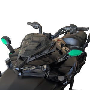 May include: Close-up of a black motorcycle with a tinted windshield and green side mirrors. The handlebars and controls are visible, along with the text "VELOCIMEDS" on the windshield. The motorcycle has a sleek, modern design.