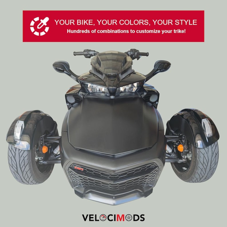 K&ouml;nnte beinhalten: Ein mattschwarzes Trike mit drei R&auml;dern, das ein schlankes Design und moderne Scheinwerfer aufweist. Das Bild enth&auml;lt den Text "YOUR BIKE, YOUR COLORS, YOUR STYLE" und "VELOCIMODS". Die Front des Trikes hat einen Wabenf&ouml;rmigen K&uuml;hlergrill.