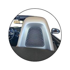 May include: A silver car headrest with a perforated black mesh insert. The headrest is part of the interior of a convertible car, with the steering wheel and side mirror visible in the background.