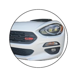 May include: White sports car front view with black grill and red "124 Spider" emblem. The car has a sleek design with a prominent headlight and fog lights. The car is in a circular frame.