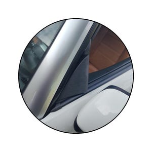 Op de afbeelding: Een close-up van de buitenkant van een auto, met een zilveren afwerking, een zwart driehoekig element en een witte zijspiegel. Het interieur toont een glimp van bruine leren bekleding.