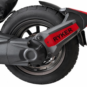May include: A black motorcycle rear wheel assembly featuring a textured tire, a dark metal rim, and a matte black swingarm component. A prominent red accent panel is attached to the side, displaying the black text RYKER in bold capital letters.