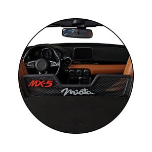 Puede incluir: Vista interior del salpicadero de un coche. El salpicadero es negro y marrón con volante, indicadores y consola central. El texto "MX-5" es rojo y "Miata" plateado.