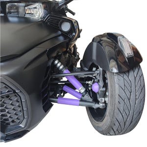 Puede incluir: Primer plano de un vehículo negro de tres ruedas, mostrando la rueda delantera y el sistema de suspensión. Los componentes de la suspensión son de un vibrante color púrpura, contrastando con la carrocería y la rueda negras. El neumático tiene un dibujo grueso.