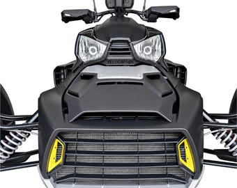 Puede incluir: Un vehículo de tres ruedas en negro mate con una rejilla frontal prominente con detalles en amarillo, faros dobles y manillar con protectores de manos. La palabra VELOCIMODS está impresa en letras negras en el panel central sobre la rejilla.