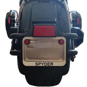 May include: Rear view of a black Spyder motorcycle. Features a red reflector, two red circular lights, and a license plate frame with the word "SPYDER" in white. The motorcycle has black fenders and a black tire.