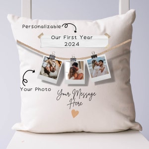 May include: Personalized throw pillow with a white background and a rope with three instant photos hanging from it. The text "Our First Year 2024" is written on a white banner above the photos. The text "Your Photo" is written below the left photo and "Your Message Here" is written below the photos with a small heart.