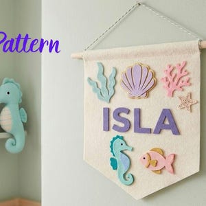 May include: A cream-colored felt wall banner featuring the name ISLA in purple letters, decorated with felt cutouts of a light blue seahorse, a pink fish, a purple seashell, a light blue seaweed sprig, a pink coral branch, and a starfish.