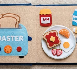 May include: A felt quiet book page featuring a light blue toaster with the word TOASTER, two slices of bread, a red jar labeled JAM, a blue and white milk carton labeled MILK, a butter stick labeled BUTTER, a plate with toast, egg, orange, and strawberries, plus a fork and spoon.