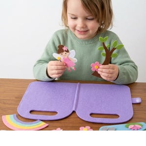Peut inclure: Un ensemble de jeu en feutrine violette comprenant un plateau pliable avec poignées, accompagné de diverses découpes en feutrine dont une fée aux ailes blanches, un arbre marron aux feuilles vertes, une fleur rose et un arc-en-ciel.