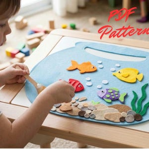 Può includere: Un progetto creativo in feltro azzurro a forma di boccia per pesci con pesci in feltro arancioni, gialli e viola, alghe verdi e sassolini marroni. Il testo PDF Pattern è stampato in rosso nell'angolo in alto a destra dell'attività.