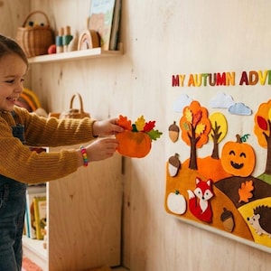 May include: A child interacts with a felt autumn-themed wall decoration, holding a felt pumpkin with leaves. The wall art features trees, a fox, a hedgehog, and the text "MY AUTUMN ADVENTURE."