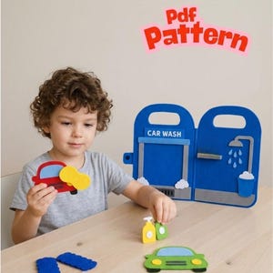 Puede incluir: Un juego de lavado de coches de fieltro que incluye una estructura plegable azul con un gráfico de estación de lavado y ducha, un coche rojo con una esponja amarilla, un coche verde, botellas pequeñas y formas de nubes blancas. El texto Pdf Pattern es visible en rosa.