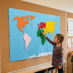Könnte beinhalten: Eine große Weltkarte aus Filz als Wandbehang mit einem leuchtend blauen Ozeanhintergrund und Kontinenten in Orange, Pink, Rot, Gelb und Grün. Die Karte ist auf einer Pinnwand neben verschiedenen Lernplakaten mit Alphabet und Zeichnungen angebracht.