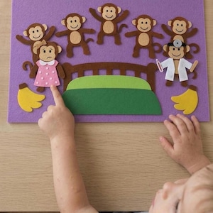 May include: A purple felt board featuring five brown monkeys, a brown bed with a green blanket, two yellow banana clusters, and a monkey dressed as a doctor with a white coat and head mirror. One monkey wears a pink dress with white polka dots.