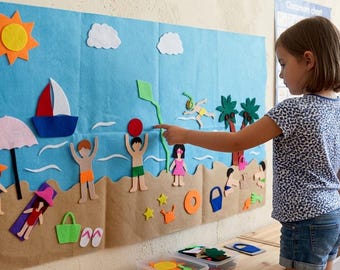 May include: A large blue felt wall hanging featuring a beach scene with tan sand, a yellow sun, white clouds, a sailboat, a pink umbrella, and various felt figures including people, palm trees, a kite, and beach accessories like a bucket and ball.