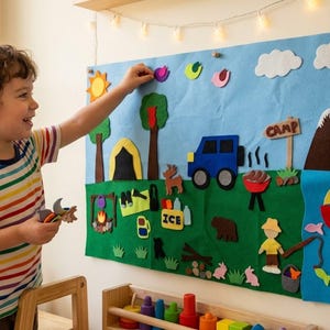 May include: A large rectangular felt board featuring a camping scene with a blue sky, white clouds, a yellow sun, green trees, a yellow tent, a blue vehicle, a brown mountain, a red boat on blue water, and various small felt shapes like fish and animals.