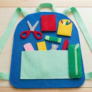 May include: A blue felt backpack featuring light green straps and a front pocket. The surface is decorated with felt school supply shapes including red scissors, a red square, a yellow crayon holder, a green glue stick, a ruler, a sharpener, and a green pencil case.