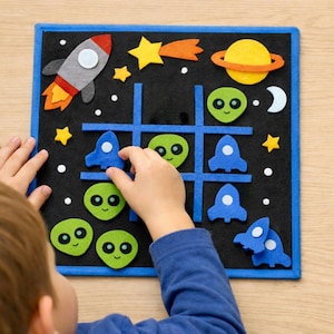 Puede incluir: Un tablero de tic-tac-toe de fieltro negro con un borde azul, con piezas de juego con temática espacial. Las piezas incluyen cohetes, extraterrestres, estrellas, un planeta y una luna creciente. Un niño está jugando.
