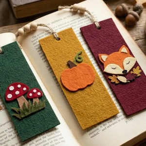 May include: Four rectangular felt bookmarks featuring autumn-themed appliqués including a brown acorn and yellow leaf, two red and white mushrooms, an orange pumpkin, and a fox face. Each bookmark has a jute twine tassel with wooden beads.