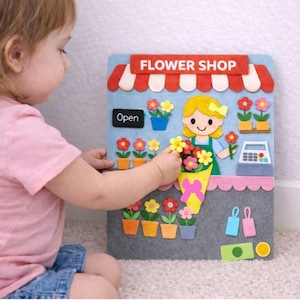 Può includere: Un set da gioco di fiorista in feltro con una tenda a strisce rosse e bianche, le parole "FLOWER SHOP" e "Open" sono visibili. Il set da gioco presenta una fioraia, fiori in vaso e un registratore di cassa. Un bambino interagisce con il set.