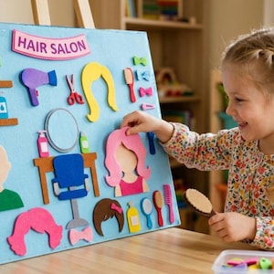 May include: A child plays with a felt board labeled "Hair Salon." The board features felt cutouts of hair styles, salon tools, and a mirror. The child is holding a brush and smiling.