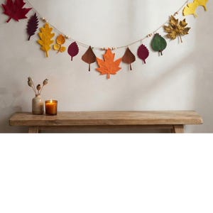 May include: A decorative garland featuring various felt leaves in shades of red, orange, yellow, brown, purple, and green, strung along a thin cord with small wooden beads interspersed between the leaf shapes to create a seasonal hanging display.
