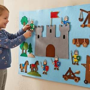 May include: A felt castle playset with a blue background. The set includes a gray castle, trees, horses, and various felt figures of knights and siege weapons. A child is interacting with the playset.