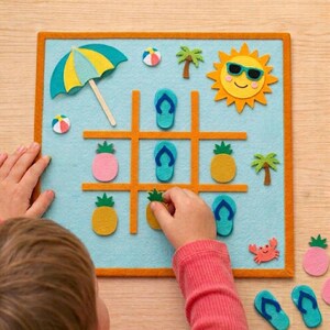 May include: A square felt tic-tac-toe game board with a light blue background and orange grid lines. The game features felt pieces including yellow pineapples, pink pineapples, and blue flip-flops. Decorative felt accents include a beach umbrella, a sun with sunglasses, palm trees, a beach ball, and a crab.