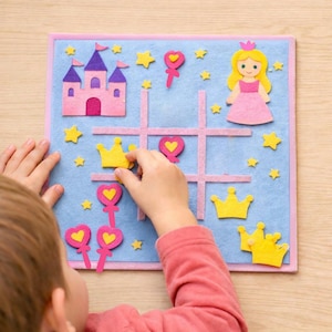 Puede incluir: Un tablero de tic-tac-toe de fieltro azul con borde rosa, con un castillo rosa, estrellas amarillas y una princesa. El juego incluye coronas amarillas y piezas de juego rosas en forma de corazón. La mano de un niño coloca una corona en el tablero.