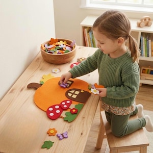 Puede incluir: Un set de manualidades de fieltro con forma de casa de calabaza que incluye una base naranja grande con puerta y ventana marrones, acompañado de varias piezas de fieltro como setas rojas, hojas verdes, flores coloridas y mariposas moradas.