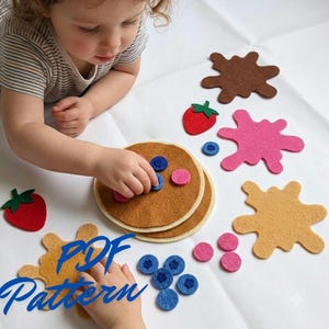 Op de afbeelding: Een vilt pannenkoeken speelset met twee ronde lichtbruine pannenkoeken, diverse vilten fruitstukjes zoals rode aardbeien en blauwe bosbessen, en kleurrijke siroopspatten in bruin, roze en beige. De afbeelding bevat de tekst PDF Pattern.