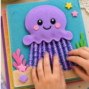 May include: A felt activity book page featuring a purple jellyfish with black eyes and pink cheeks. The jellyfish has dangling purple beaded tentacles. The background is blue with felt seaweed, a seashell, and small yellow and pink stars.