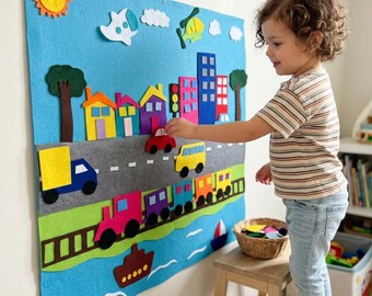 May include: A large blue felt wall hanging featuring a colorful city scene with houses, buildings, a gray road with vehicles, a train on tracks, a boat on water, a yellow sun, and clouds. The felt pieces are removable and arranged on the board.