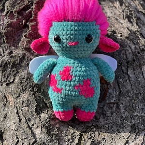 May include: A crocheted plush toy with a pink fuzzy hair, blue body, pink accents, and white wings. The toy is sitting on a tree trunk.