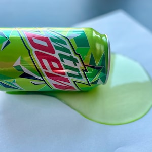 May include: A green and red Mountain Dew can is lying on its side on a white surface. The can is leaking green liquid, forming a puddle around it.