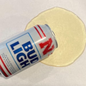 May include: A silver and blue Bud Light beer can lies on its side next to a spill of a light yellow liquid. The can has red accents and the words "Bud Light" in blue.