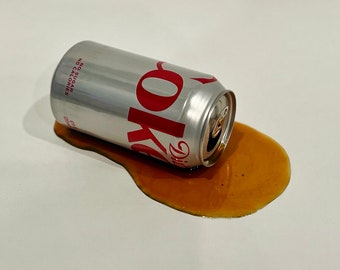 May include: A silver can of Diet Coke with the words "Diet Coke" and "No Sugar, Calories" printed on the side, lying on a white surface with a brown liquid spilled around it.