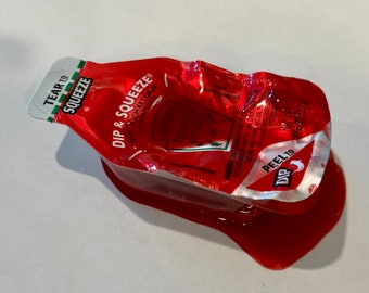 May include: A partially used red ketchup packet with the text "DIP & SQUEEZE" and "TEAR TO SQUEEZE". Ketchup is spilled out of the packet. The net weight is 0.95 oz (27g).