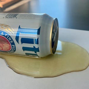 May include: A can of Miller Lite beer has spilled on a white surface, creating a puddle of golden liquid. The can is lying on its side, with the Miller Lite logo and branding visible.