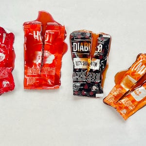 May include: Four different types of hot sauce packets, all of which have been squeezed and are now empty. The packets are on a white surface. The sauces are "Fire!" "Hot!" "Diablo" and "Mild".